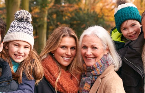 A smiling family with an autumnal woodland background. The family are made up of parents, grandparents and a young boy and girl. They are all wearing warm winter cloths and look to be happy and enjoying time together.