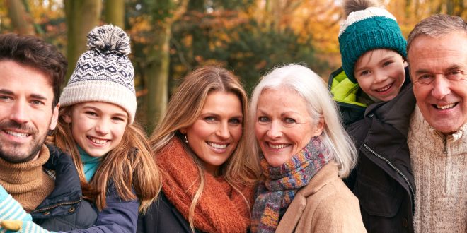 A smiling family with an autumnal woodland background. The family are made up of parents, grandparents and a young boy and girl. They are all wearing warm winter cloths and look to be happy and enjoying time together.