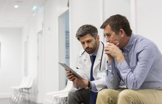 A doctor holding a digital tablet is talking with a middle aged man. They are sitting in a hospital corridor.