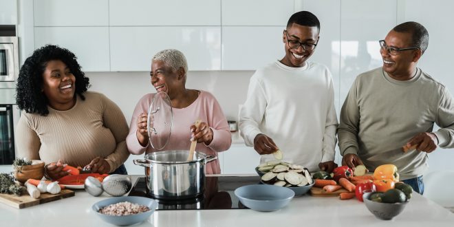 A family group are are cooking together in a modern looking kitchen. From left to right they are a young black woman, she is smiling at an older black woman who is stirring a large cooking pot. A young black man wearing glasses and an older black man wearing glasses are also smiling and helping to prepare colourful vegetables.