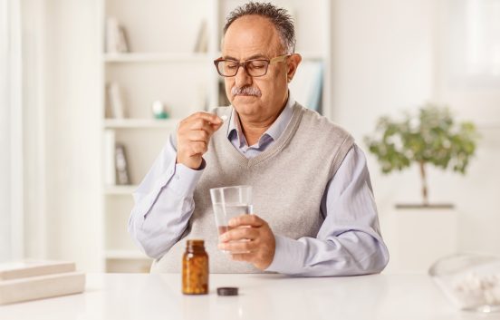 An older man is sat at a table. He has a bottle of tablets in front of him. He is looking thoughtfully at one of the tablets in his hand. In his other hand he holds a glass of water.