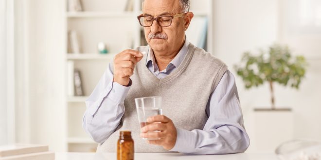 An older man is sat at a table. He has a bottle of tablets in front of him. He is looking thoughtfully at one of the tablets in his hand. In his other hand he holds a glass of water.