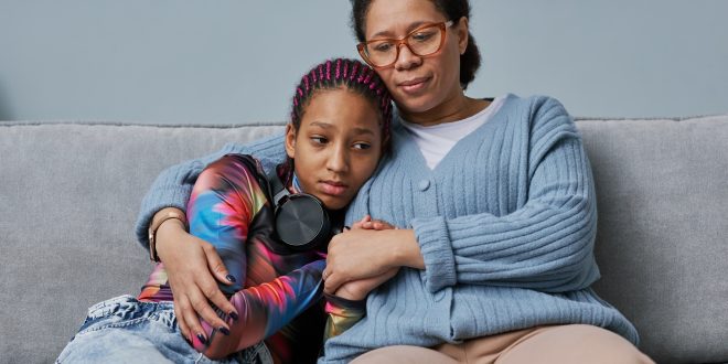 A mum hugs her teenage daughter who looks worried. The are sitting on a sofa.