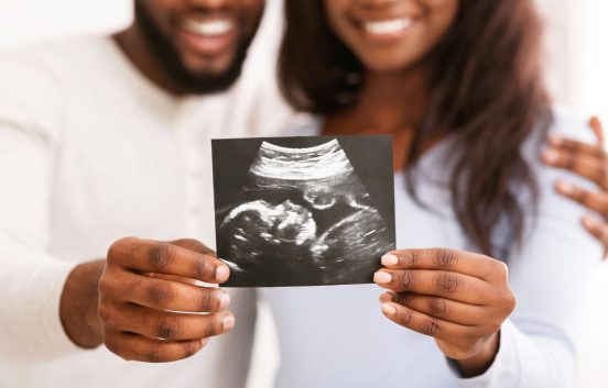 A young black couple are holding an ultrasound picture from a pregnancy scan. We only see the bottom part of the couple's faces. The man has a short beard, the woman has long wavey hair. They are both wearing white long sleeved tops.