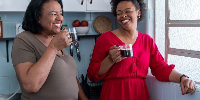 Two black women are drinking coffee from glass mugs. They are smiling and standing in a kitchen next to a window. From their ages they could be mother and daughter.